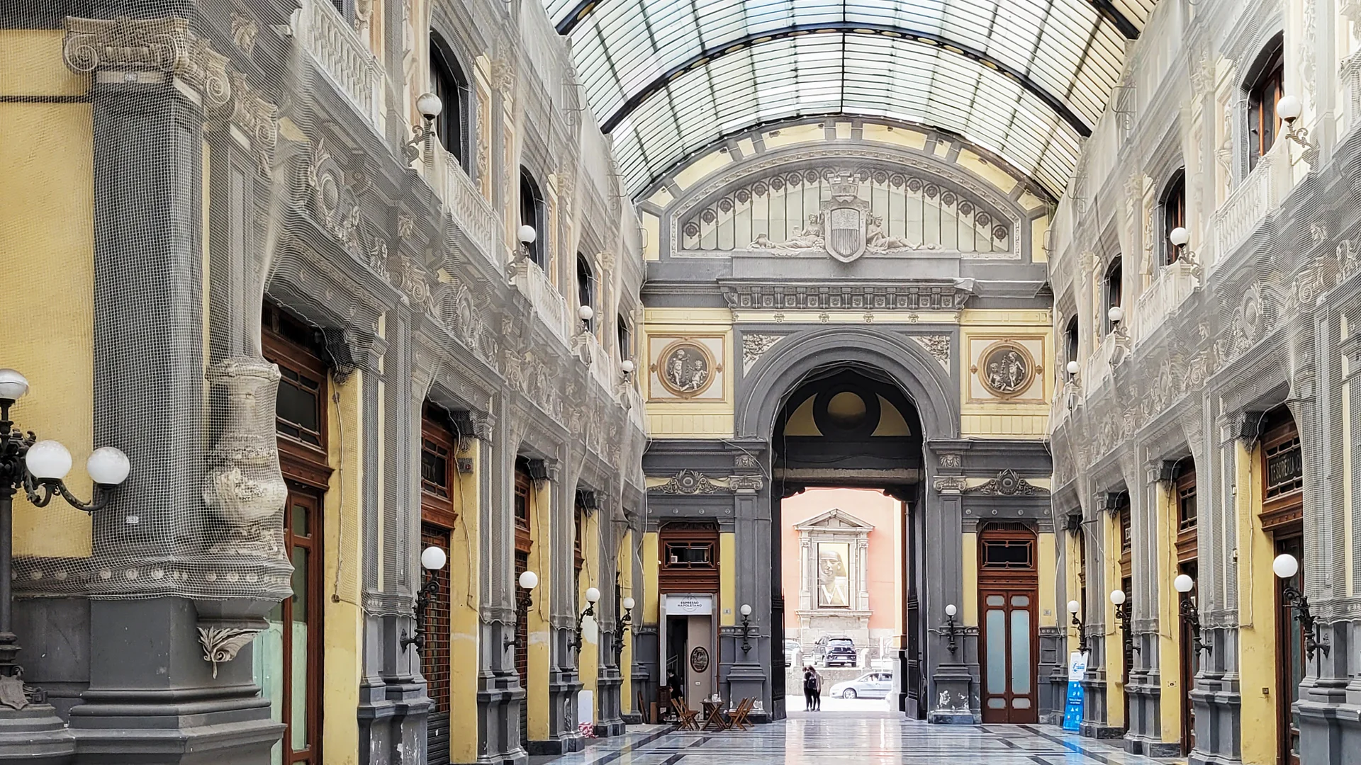 Inside the Galleria Principe di Napoli: a long glass-and-iron vaulted arcade with painted yellow and grey facades and a marble floor